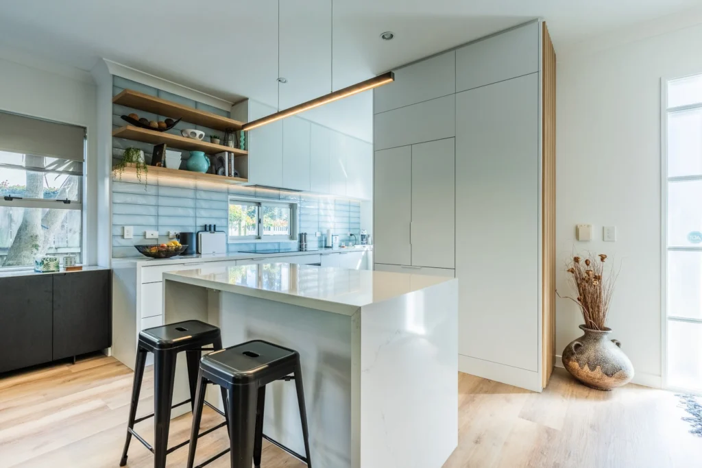 Modern minimalist kitchen with a white island, black bar stools, open wooden shelves, and a hanging linear light fixture