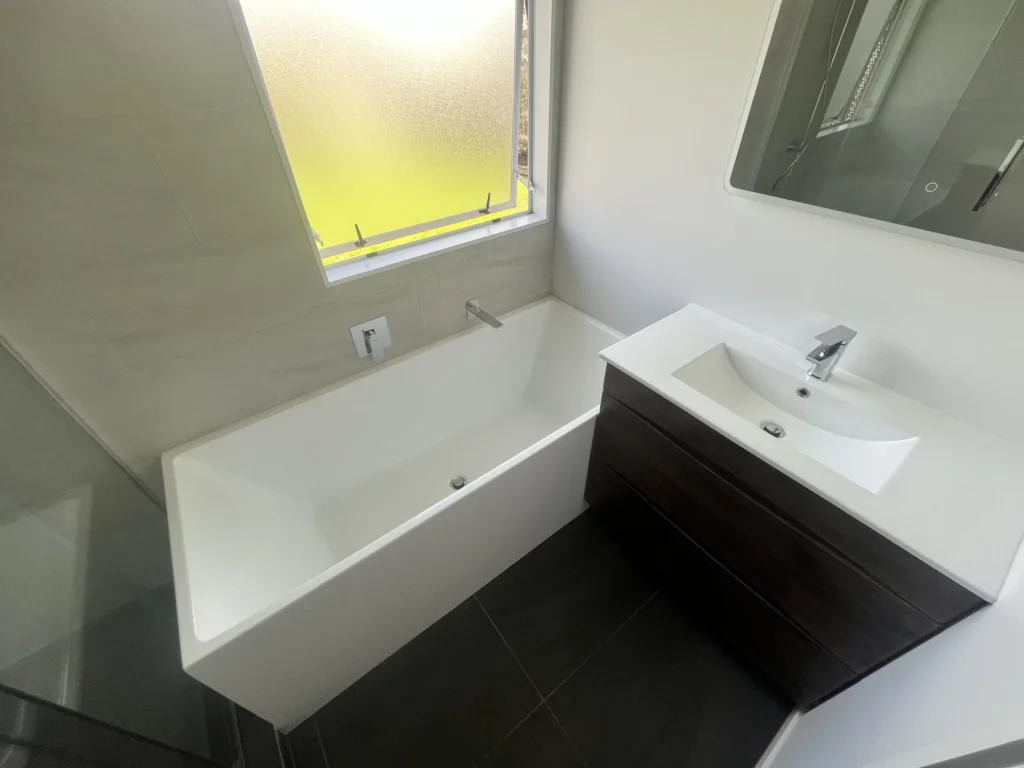 Modern bathroom with a white bathtub, wall-mounted faucet, vanity sink, and frosted window.